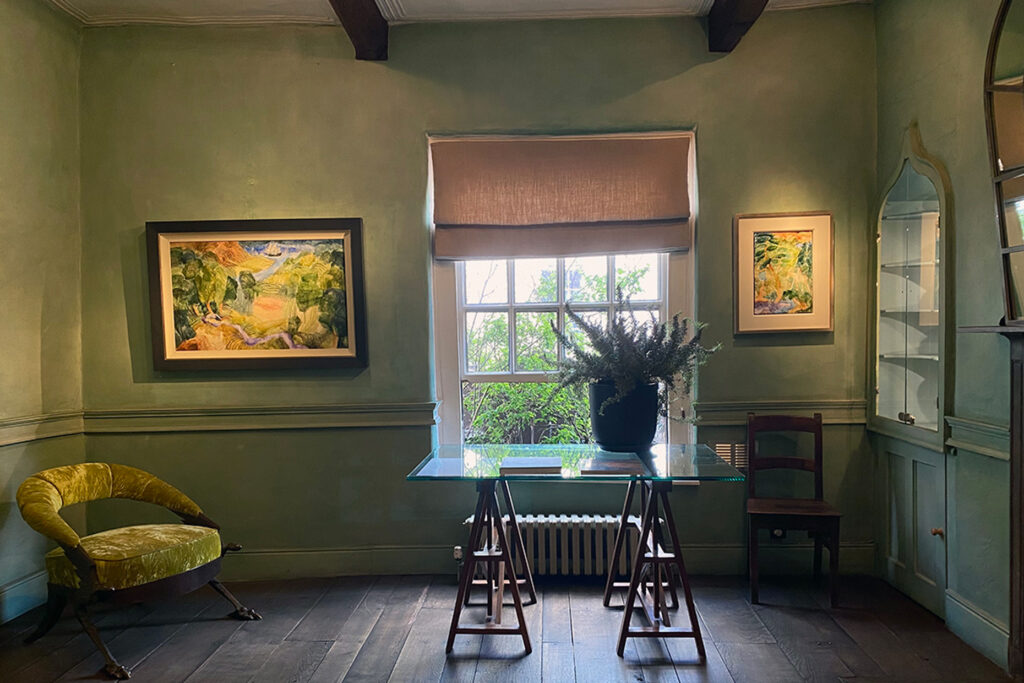 Upstairs at The Art Shop on Cross Street. Sage-green panelled walls with two framed Eleri Mills landscape paintings either side of a tall sash window, a soft pink linen blind half-down. A glass-topped trestle table in the middle of the room holds art books. A moss-green velvet tub chair sits in the corner. Dark-stained timber floor, glazed corner display cupboard.